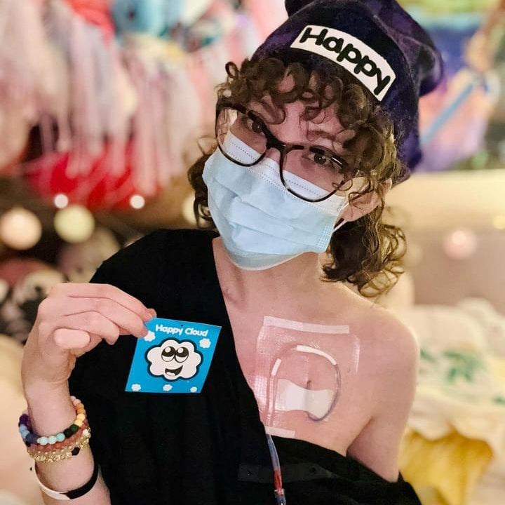 Person wearing a face mask and 'Happy' beanie holding a 'Happy Cloud' card in a colorful setting.